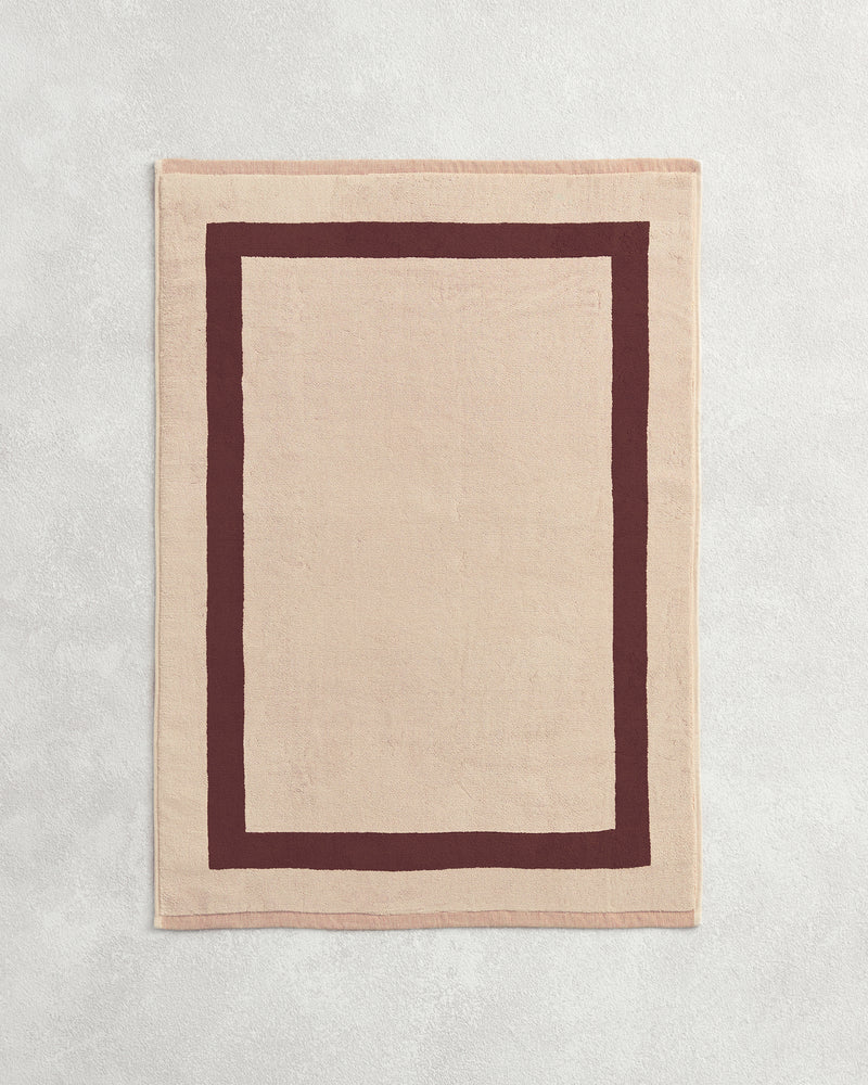 Flat cotton terry bath mat on a grey background in a creme colour with a cacao border.
