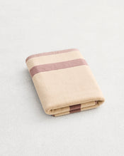 A folded cotton terry bath mat on a grey background, in a creme colourway with a lavender border.  - view