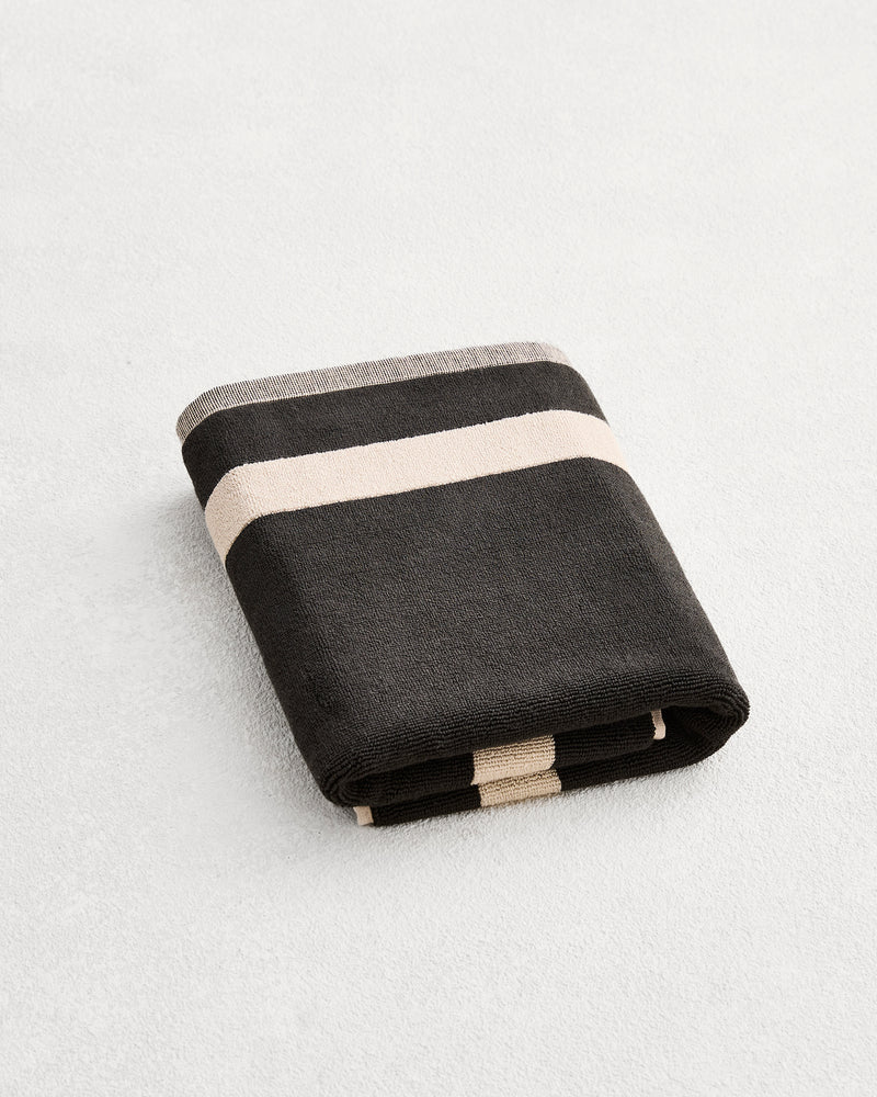 A folded cotton terry bath mat on a grey background, in a charcoal colourway with an oatmeal border. .