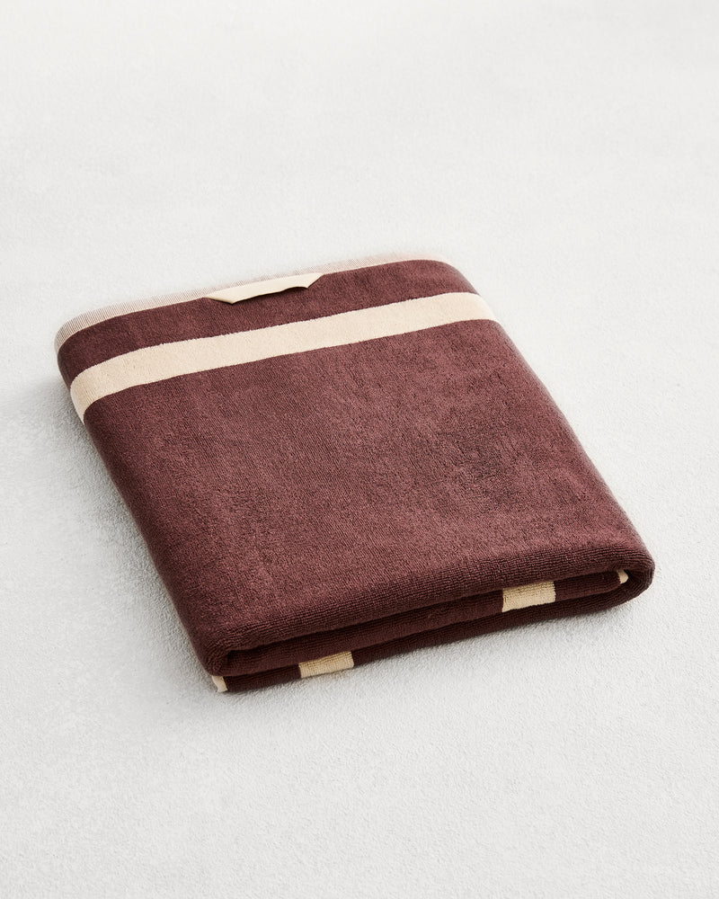 A folded cotton terry bath sheet on a grey background, in a cacao colourway with a creme border. 
