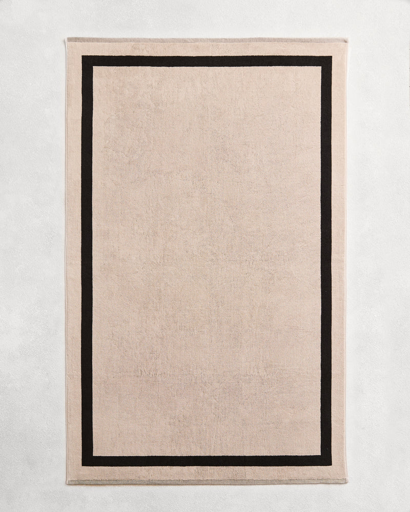 Flat cotton terry bath towel on a grey background in an oatmeal colour with a charcoal border.