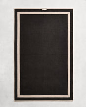 Flat cotton terry bath towel on a grey background in a charcoal colour with an oatmeal border. - view