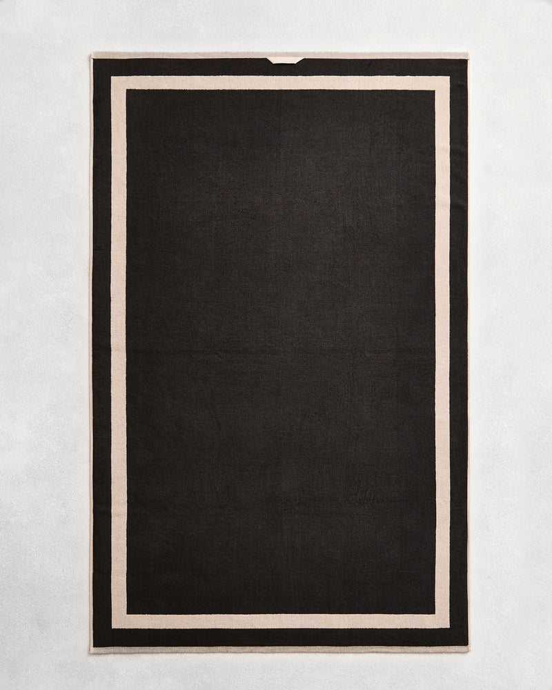 Flat cotton terry bath towel on a grey background in a charcoal colour with an oatmeal border.