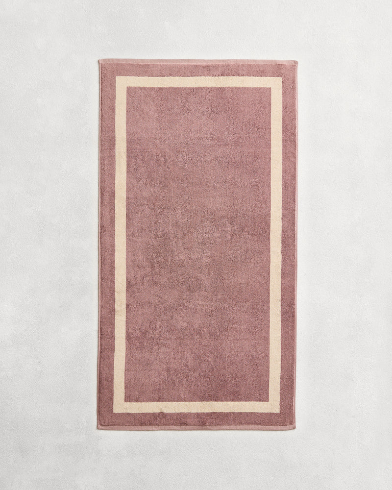 Flat cotton terry bath towel on a grey background in a lavender colour with a creme border.