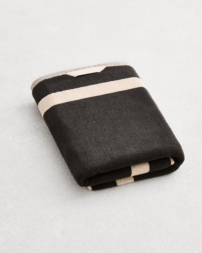 A folded cotton terry bath towel on a grey background, in a charcoal colourway with an oatmeal border. .