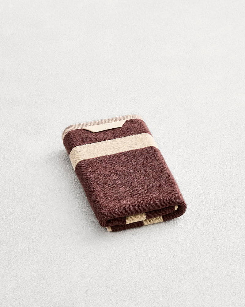 A folded cotton terry hand towel on a grey background, in a cacao colourway with a creme border. 