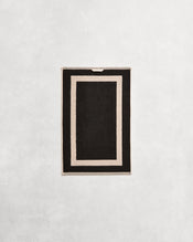Flat cotton terry bath towel on a grey background in a charcoal colour with an oatmeal border. - view