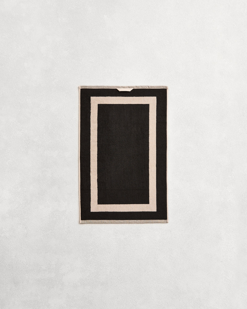 Flat cotton terry bath towel on a grey background in a charcoal colour with an oatmeal border.