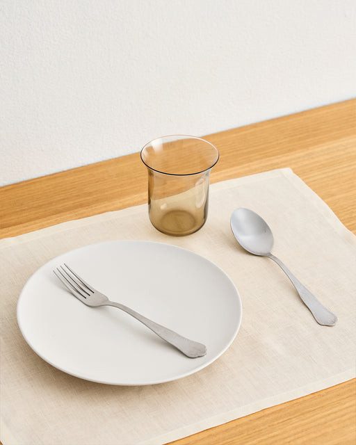A simple table setting with a white plate, cutlery, and glass—all arranged on Bed Threads’ Crème 100% French Flax Linen Placemats (Set of Four) for stylish protection atop a wooden table.