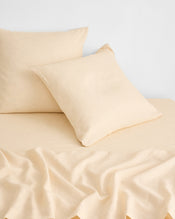 Two beige pillows rest on a matching beige bed sheet, with part of the sheet softly wrinkled in the foreground, all set against a plain white wall background. - view