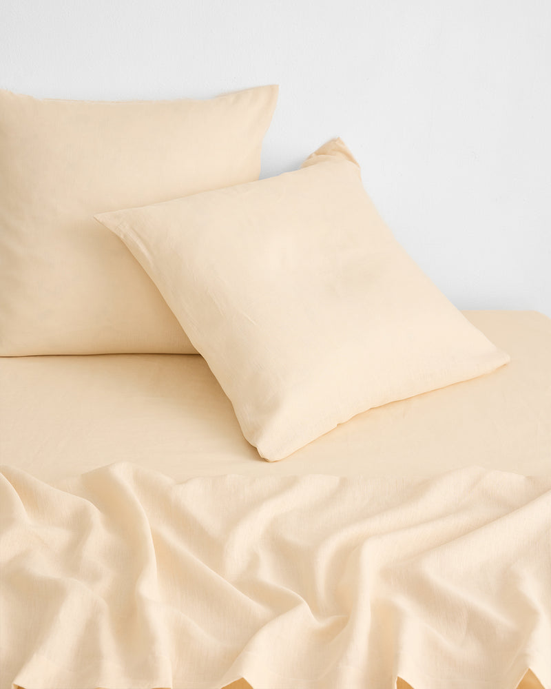 Two beige pillows rest on a matching beige bed sheet, with part of the sheet softly wrinkled in the foreground, all set against a plain white wall background.