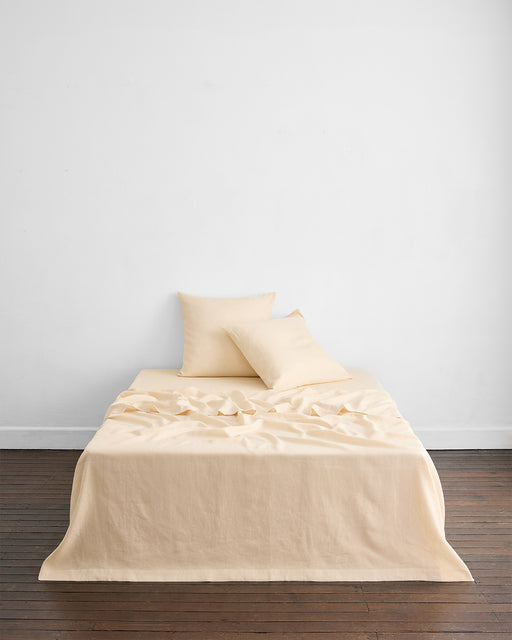 A neatly made bed with cream-colored sheets and two pillows sits against a plain white wall on a wooden floor; the bedding looks soft with some gentle wrinkles.