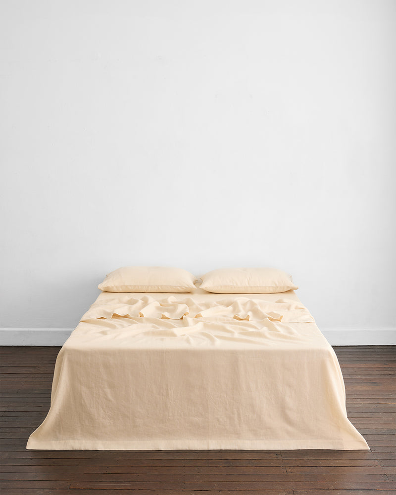 A neatly made bed with beige sheets and two pillows stands against a plain white wall on a wooden floor. The setting is minimal and uncluttered.
