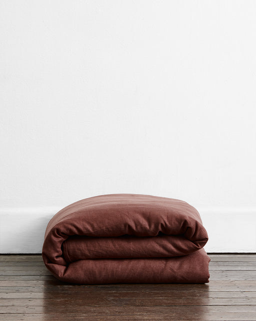 The Bed Threads Cacao & Hazelnut 100% French Flax Linen Bedding Bundle rests folded on a wooden floor by a white wall, offering stylish, temperature-regulating comfort.