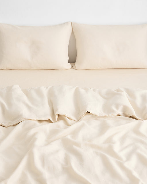 A bed neatly made with cream-colored sheets and two matching pillows, topped with the Bed Threads Crème 100% French Flax Linen Duvet Cover, slightly rumpled at the center and bottom, set against a plain white wall.
