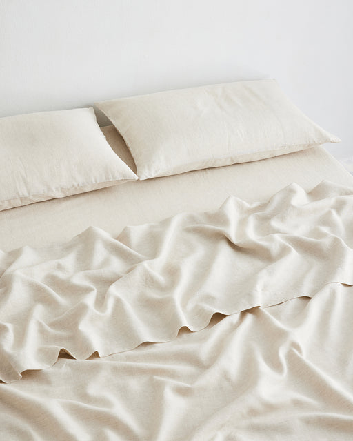 A neatly made bed with two matching beige pillows and a slightly rumpled Bed Threads Oatmeal & White Stripe 100% French Flax Linen Flat Sheet, prized for its temperature-regulating properties, sits against a plain white wall.