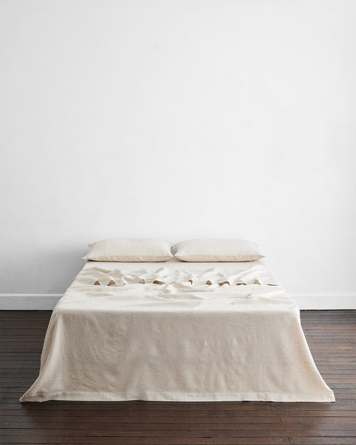 A neatly made bed with two pillows and a cream, temperature-regulating blanket is topped with the Oatmeal & White Stripe 100% French Flax Linen Flat Sheet by Bed Threads, set against a white wall on a dark wooden floor.