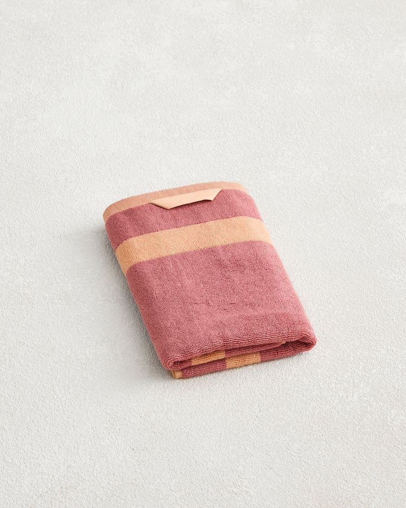 A folded Bed Threads Terracotta & Pink Clay Two-Toned 100% Cotton Terry Hand Towel with horizontal stripes rests on a light textured surface.