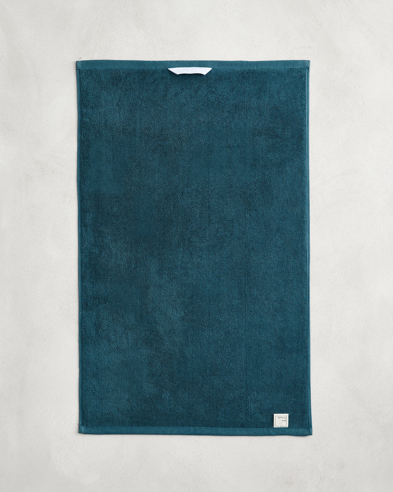 The Bed Threads Aegean 100% Cotton Terry Hand Towel (650GSM) in teal is shown flat on a light gray surface, featuring a small white label in one corner and a central hanging loop. Quick-drying and stylish for easy access.