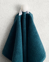 The Aegean 100% Cotton Terry Hand Towel by Bed Threads hangs on a white hook against a textured wall, folded in layers and gathered at the top with a white loop for quick drying. - view