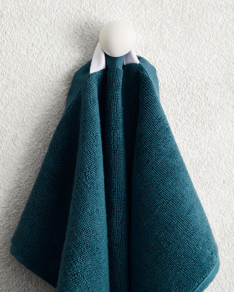 The Aegean 100% Cotton Terry Hand Towel by Bed Threads hangs on a white hook against a textured wall, folded in layers and gathered at the top with a white loop for quick drying.