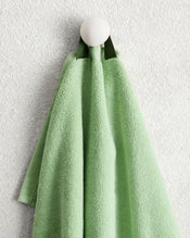 A light green Apple 100% Cotton Terry Hand Towel from Bed Threads, featuring a dark green loop, hangs on a round white wall hook against a textured white wall. - view