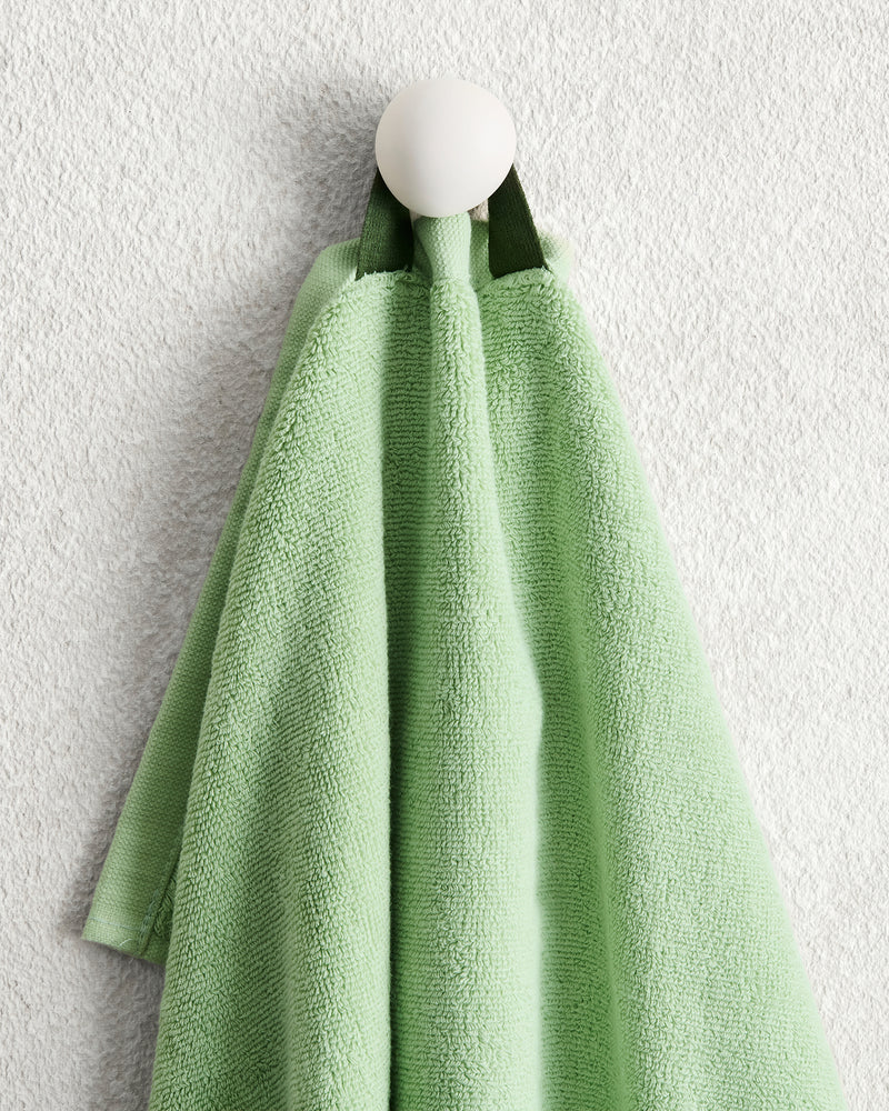 A light green Apple 100% Cotton Terry Hand Towel from Bed Threads, featuring a dark green loop, hangs on a round white wall hook against a textured white wall.