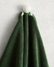 A Moss 100% Cotton Terry Hand Towel from Bed Threads hangs neatly on a round white hook, set against a textured light gray wall. - view