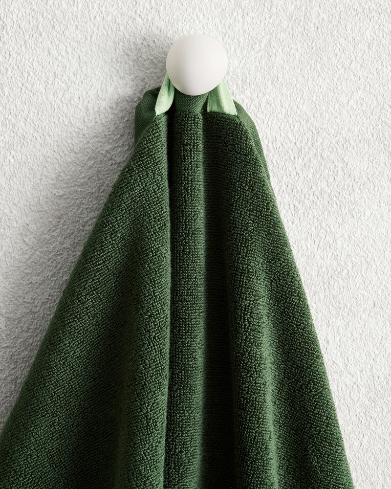 A Moss 100% Cotton Terry Hand Towel from Bed Threads hangs neatly on a round white hook, set against a textured light gray wall.