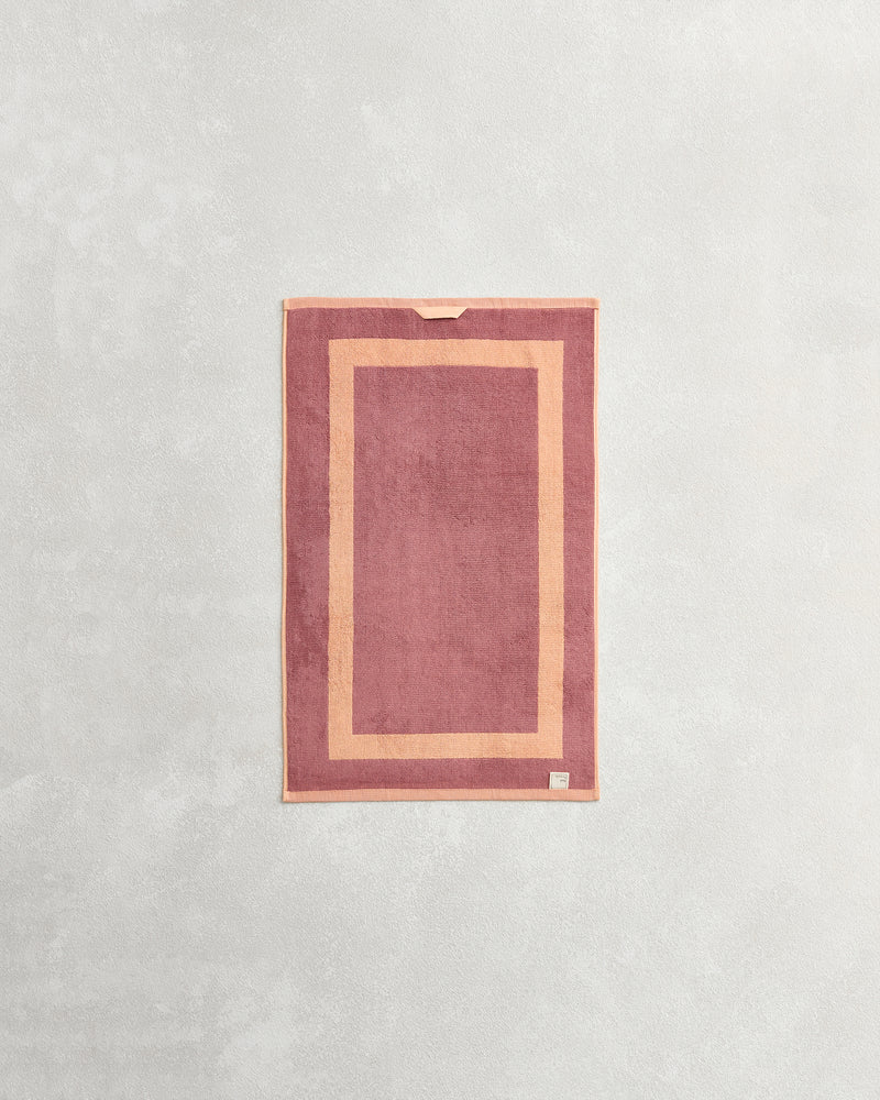 The Bed Threads Terracotta & Pink Clay Two-Toned 100% Cotton Terry Hand Towel, made from plush 650GSM quick-drying terry, features a lighter pink border, small fabric loop, and tag at the bottom corner.