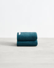 A set of two neatly folded dark teal Aegean 100% Cotton Terry Hand Towels by Bed Threads, featuring a white tag, rests on a light gray surface against a plain white background. - view