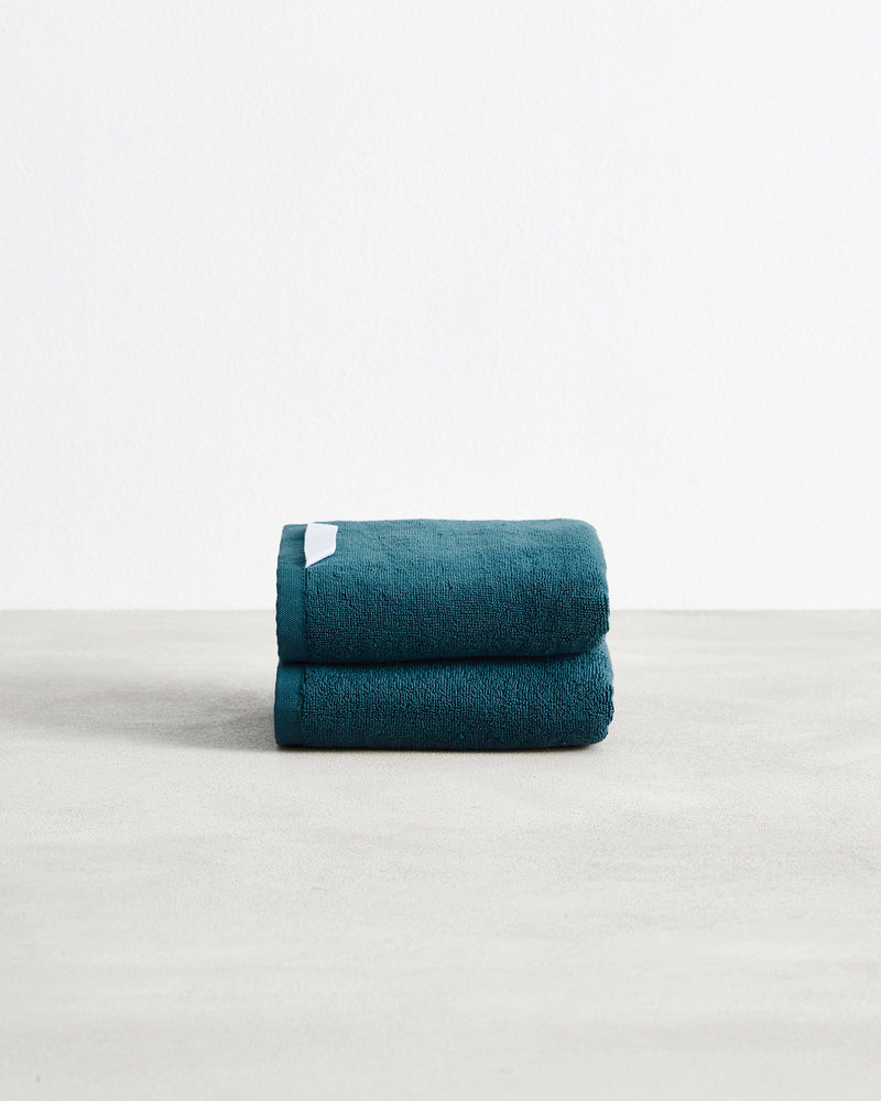 A set of two neatly folded dark teal Aegean 100% Cotton Terry Hand Towels by Bed Threads, featuring a white tag, rests on a light gray surface against a plain white background.