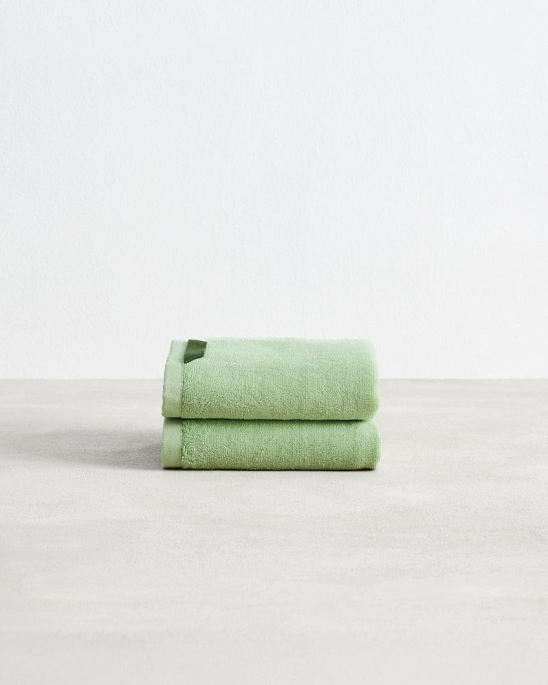 Two light green Apple 100% Cotton Terry Hand Towels by Bed Threads are neatly folded and stacked on a smooth, light surface with a plain white background.
