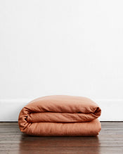 A Hazelnut & Pink Clay 100% French Flax Linen Bedding Bundle by Bed Threads is folded on a wooden floor against a plain white wall. - view