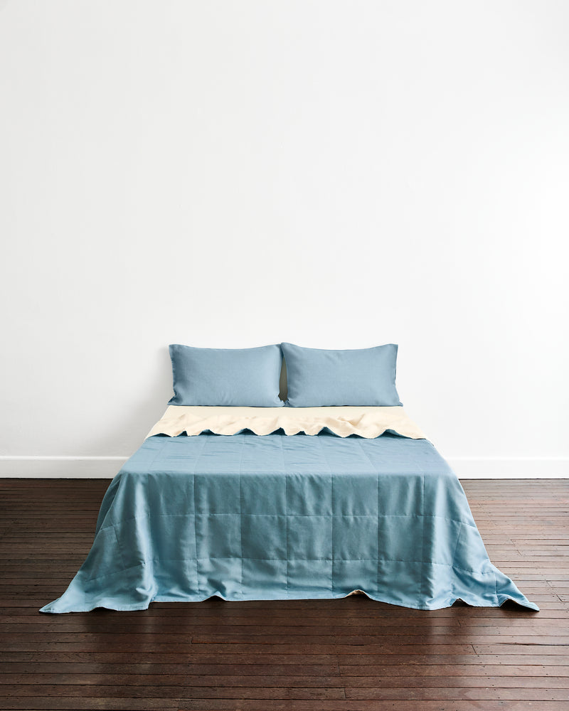 A bed dressed in a dusty blue quilt cover in Lagoon & Creme with two matching pillows in Lagoon, set against a plain white wall and dark wood floor.