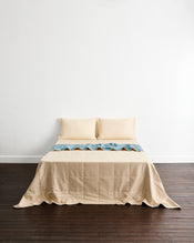 A neatly made bed with a beige quilt cover in Lagoon & Creme and two matching pillowcases in Creme, set against a plain white wall and dark wood flooring. - view