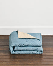 A folded dusty blue quilt cover in Lagoon & Creme with a creme underside, displayed on a dark wooden floor against a white wall. - view