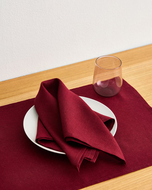 A white plate with a Bed Threads Mulberry 100% French Flax Linen Napkin sits on a matching burgundy placemat atop a wooden table, beside an empty clear glass with a hint of purple.