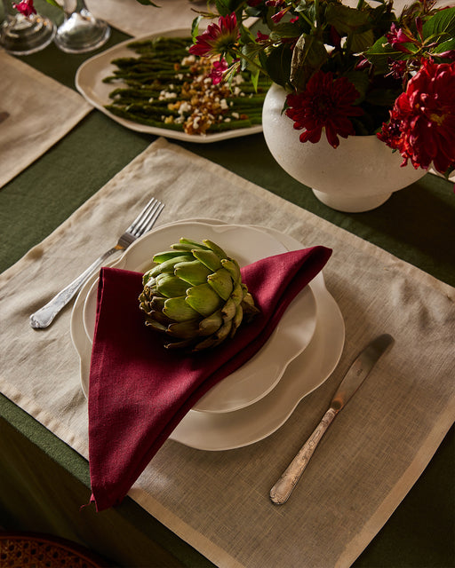 A white plate, red napkin, and artichoke rest on Bed Threads Crème 100% French Flax Linen Placemats (Set of Four), with a silver fork and knife beside the plate, plus flowers in a white vase and green beans in the background.