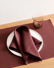 A white plate with a folded Bed Threads Cacao 100% French Flax Linen Napkin rests on a matching placemat atop a wooden table, with a transparent brown glass placed beside the plate. - view
