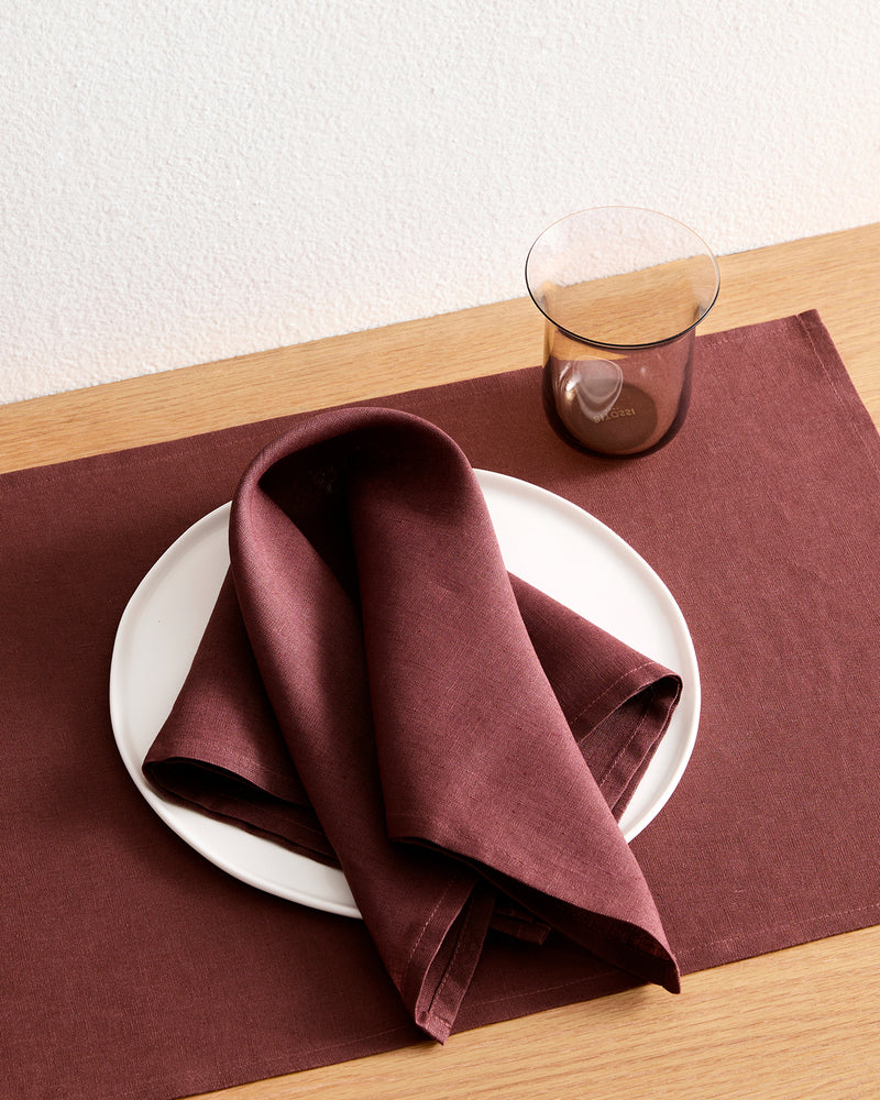 A white plate with a folded Bed Threads Cacao 100% French Flax Linen Napkin rests on a matching placemat atop a wooden table, with a transparent brown glass placed beside the plate.