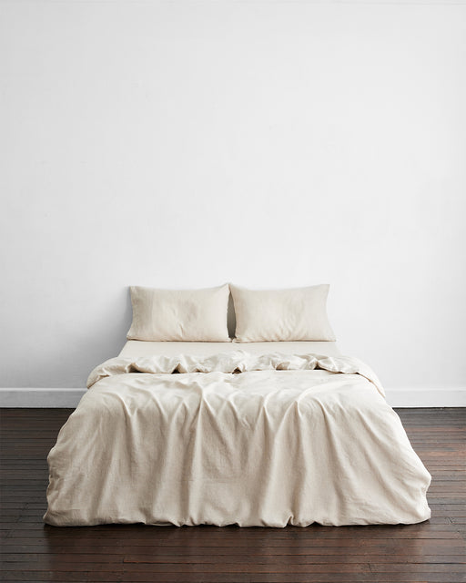 A neatly made bed with beige linens and two Bed Threads Oatmeal & White Stripe 100% French Flax Linen Pillowcases sits against a plain white wall on a dark wooden floor in a minimalist bedroom.