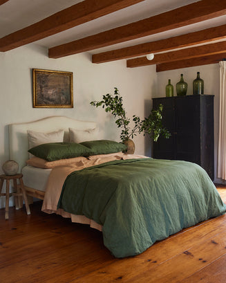 A cozy bedroom with wooden floors and exposed beams features a bed dressed in green and beige, Bed Threads Olive Stripe 100% French Flax Linen King Pillowcases for natural comfort, white pillows, a small stool, black cabinet, plant, and framed art.