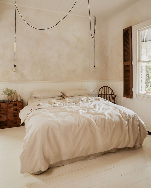 A cozy, minimal bedroom features a Bed Threads Oatmeal & White Stripe 100% French Flax Linen Flat Sheet on a double bed, pendant bulbs, a wooden chair and chest of drawers, and natural light streaming through open wooden shutters.