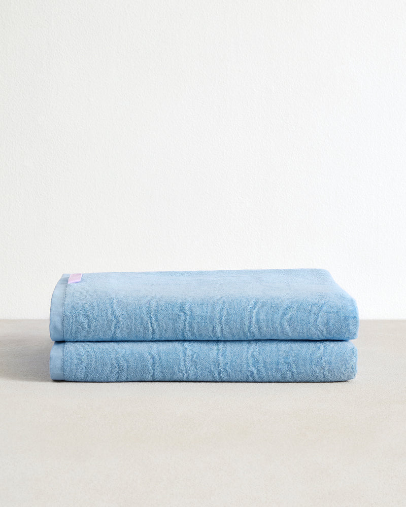 A set of two Sky 100% Cotton Terry Bath Sheets from Bed Threads are neatly folded and stacked on a light beige surface, set against a plain white background.