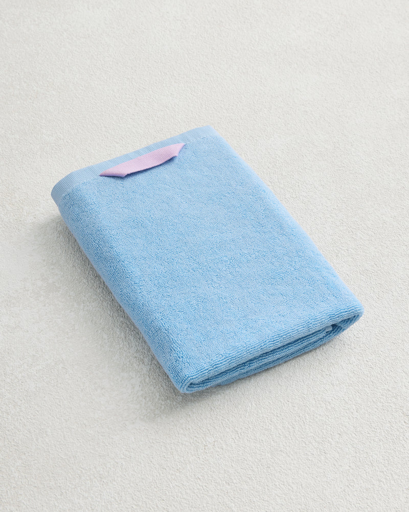 The Bed Threads Sky 100% Cotton Terry Hand Towel, light blue with a plush 650GSM texture and a small pink tag, is neatly folded on a light textured surface.