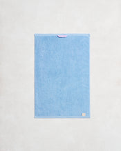 The Sky 100% Cotton Terry Hand Towel by Bed Threads hangs flat against a light wall. Plush 650GSM terry offers extra softness, while a purple loop on top and a beige label on the lower right add detail. - view