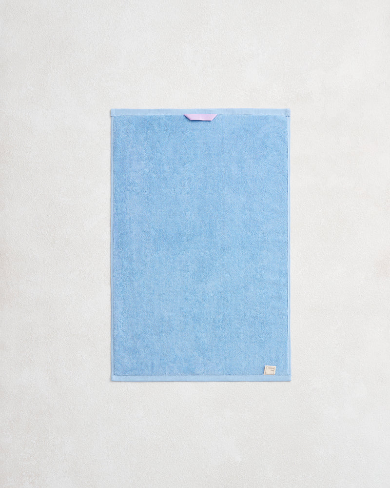 The Sky 100% Cotton Terry Hand Towel by Bed Threads hangs flat against a light wall. Plush 650GSM terry offers extra softness, while a purple loop on top and a beige label on the lower right add detail.