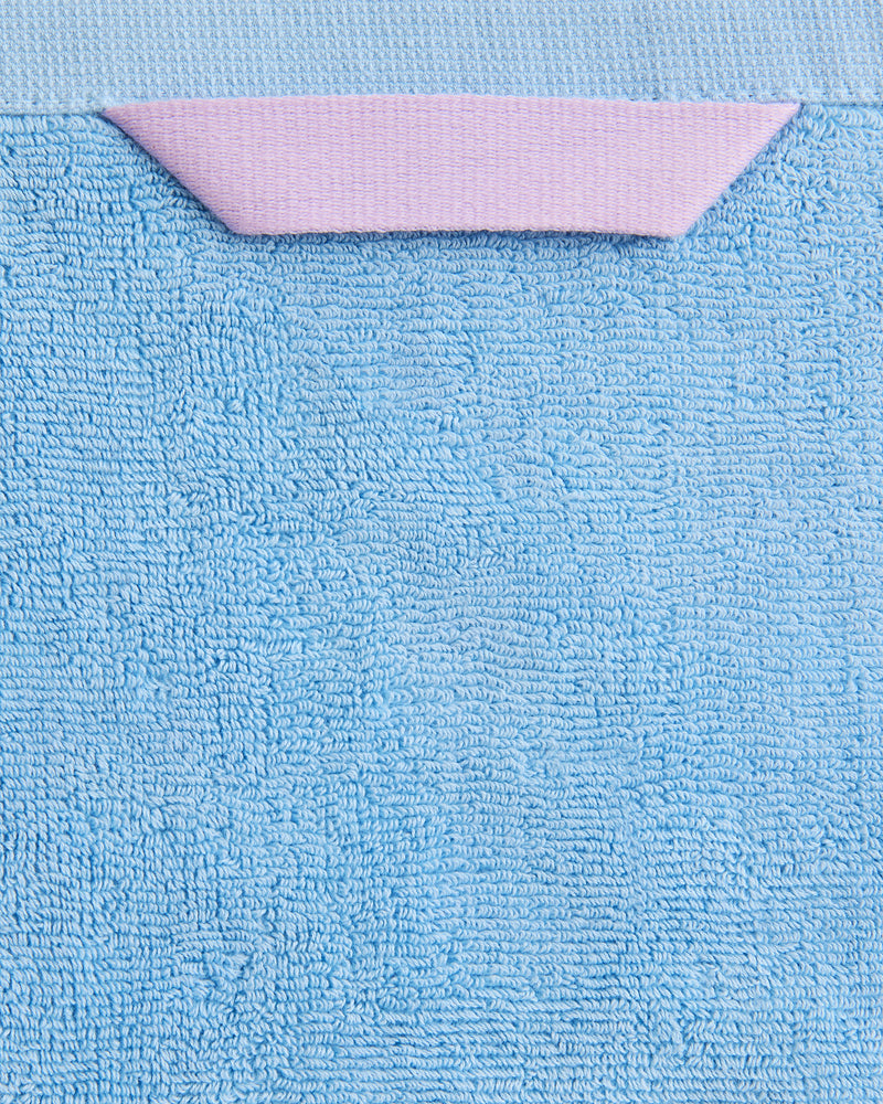 Close-up of the Bed Threads Sky 100% Cotton Terry Hand Towel (650GSM) in light blue, with a small folded pink tab at the top edge, set against a matching light blue background.