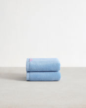 A set of two Sky 100% Cotton Terry Hand Towels by Bed Threads, in light blue, are neatly folded and stacked on a neutral surface with a white textured wall background. - view
